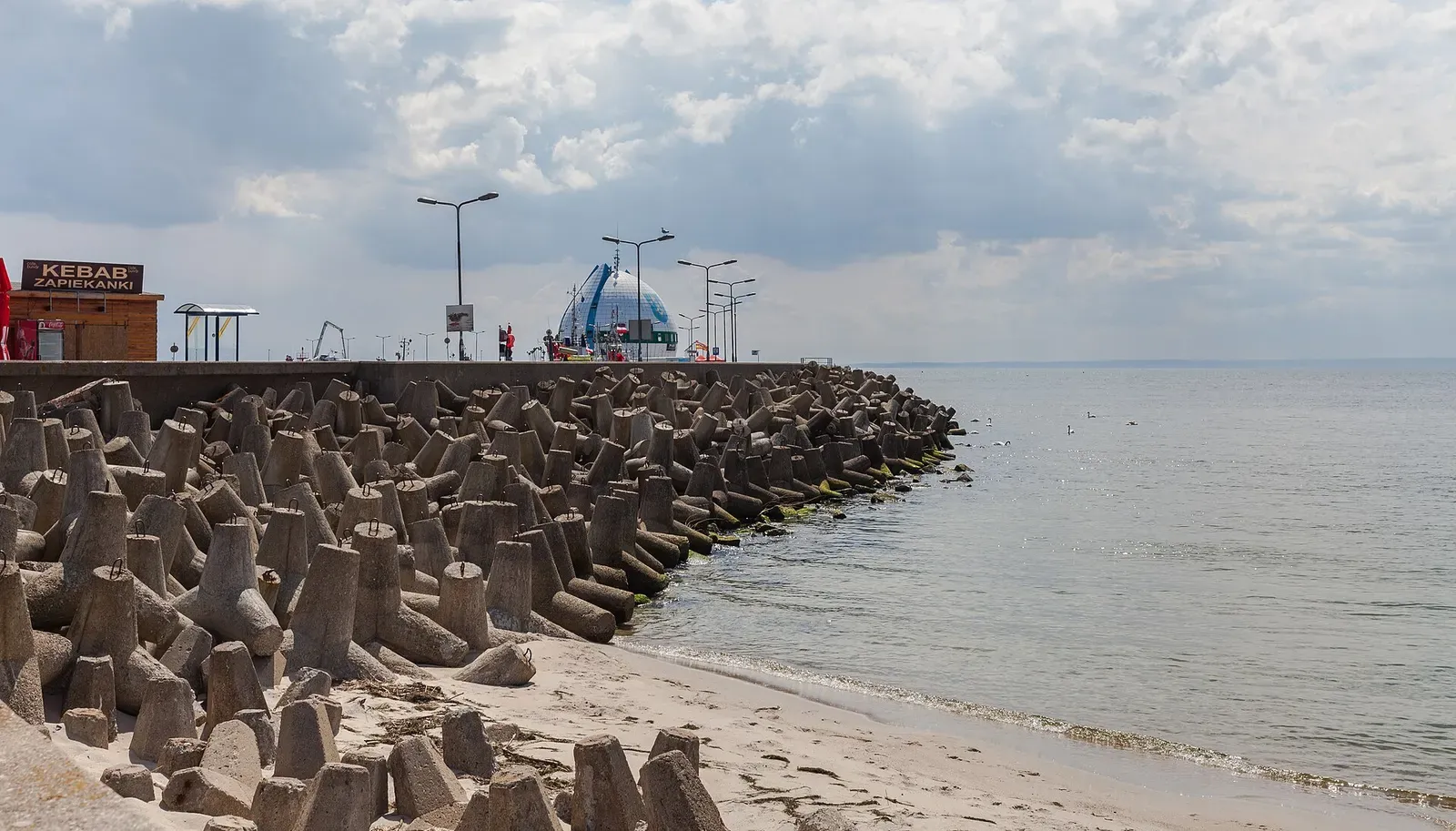 Rows of concrete tetrapods line a breakwater running out into calm grey sea beside a sandy shore, with a pier and lamp posts behind — Hel Peninsula