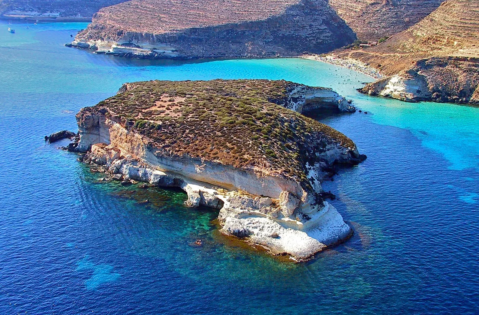 Aerial view of a low rocky islet with pale cliffs in deep blue and turquoise water, a larger island coast behind it — Lampedusa & Linosa