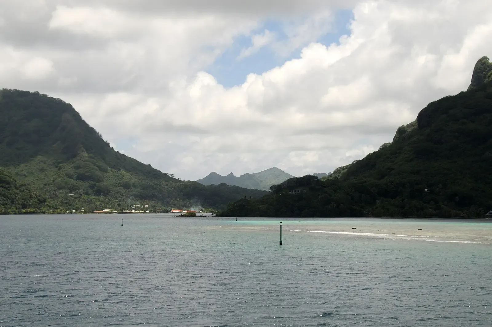 Calm grey-green water with channel markers leading toward a gap between two steep forested ridges under heavy cloud — Huahine