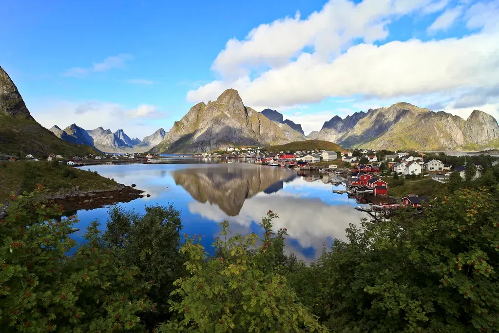 Still bay mirroring jagged grey peaks, with red and white houses along the shore and green shrubs in the foreground — Lofoten Islands
