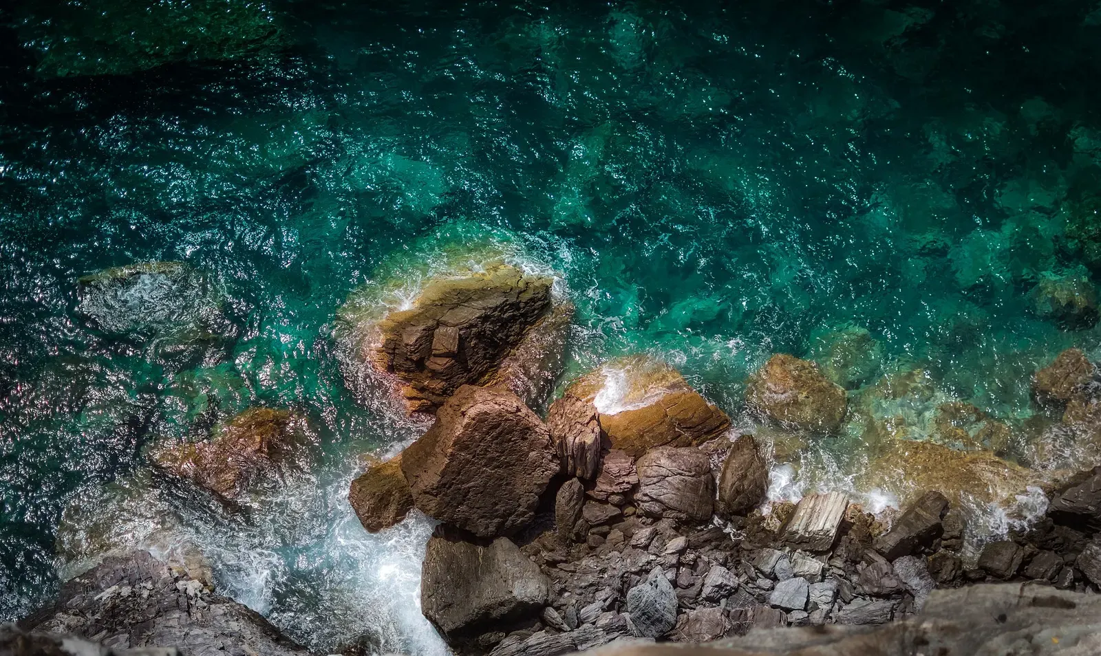 Looking straight down on brown boulders at the shoreline, with small waves breaking over them in clear green-blue water — Skopelos.