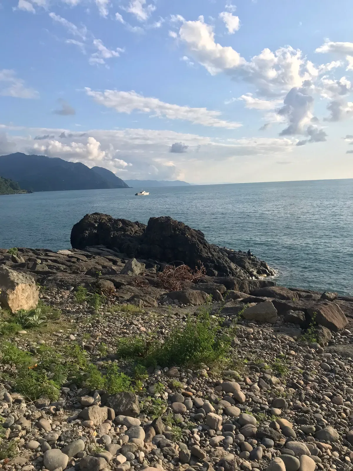 Dark rocks and a pebble shore at the edge of calm blue sea, with a small white boat offshore and forested mountains on the left — Batumi & the Adjarian Coast