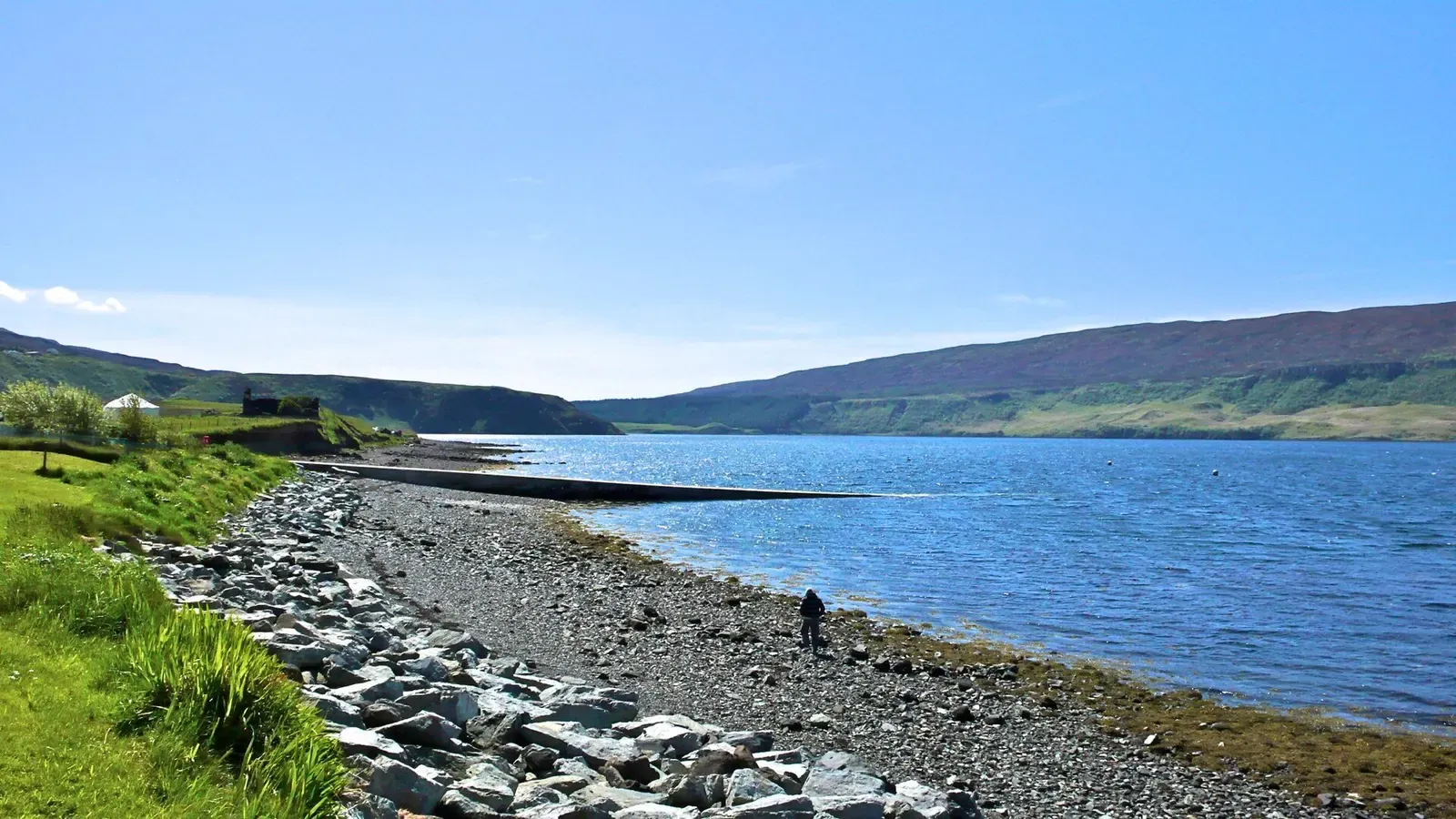 A stony shore and a long slipway run out into a bright blue loch, with one person walking the beach and low hills beyond — Isle of Skye