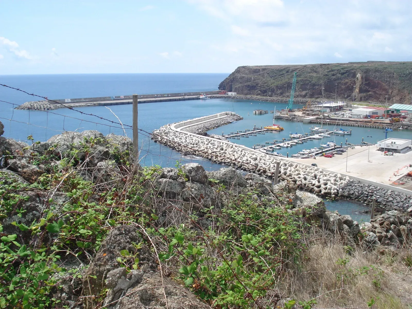 A rocky breakwater shelters a small marina with moored boats below a green headland, seen from a scrubby hillside — Azores