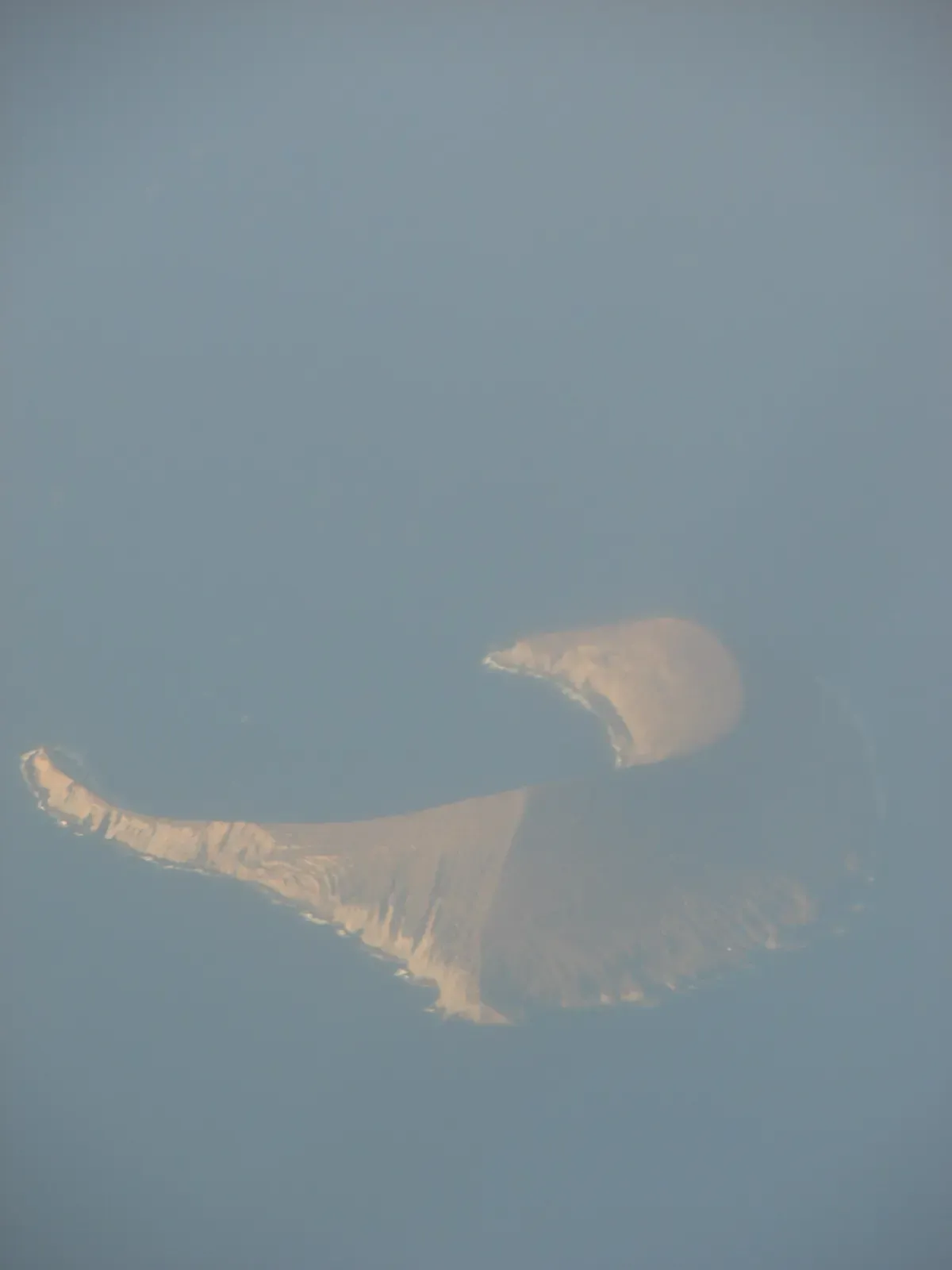 Aerial view through haze of a crescent-shaped brown volcanic islet with a long curving rim, surrounded by blue-grey sea — Vertical Awareness, Kauaʻi