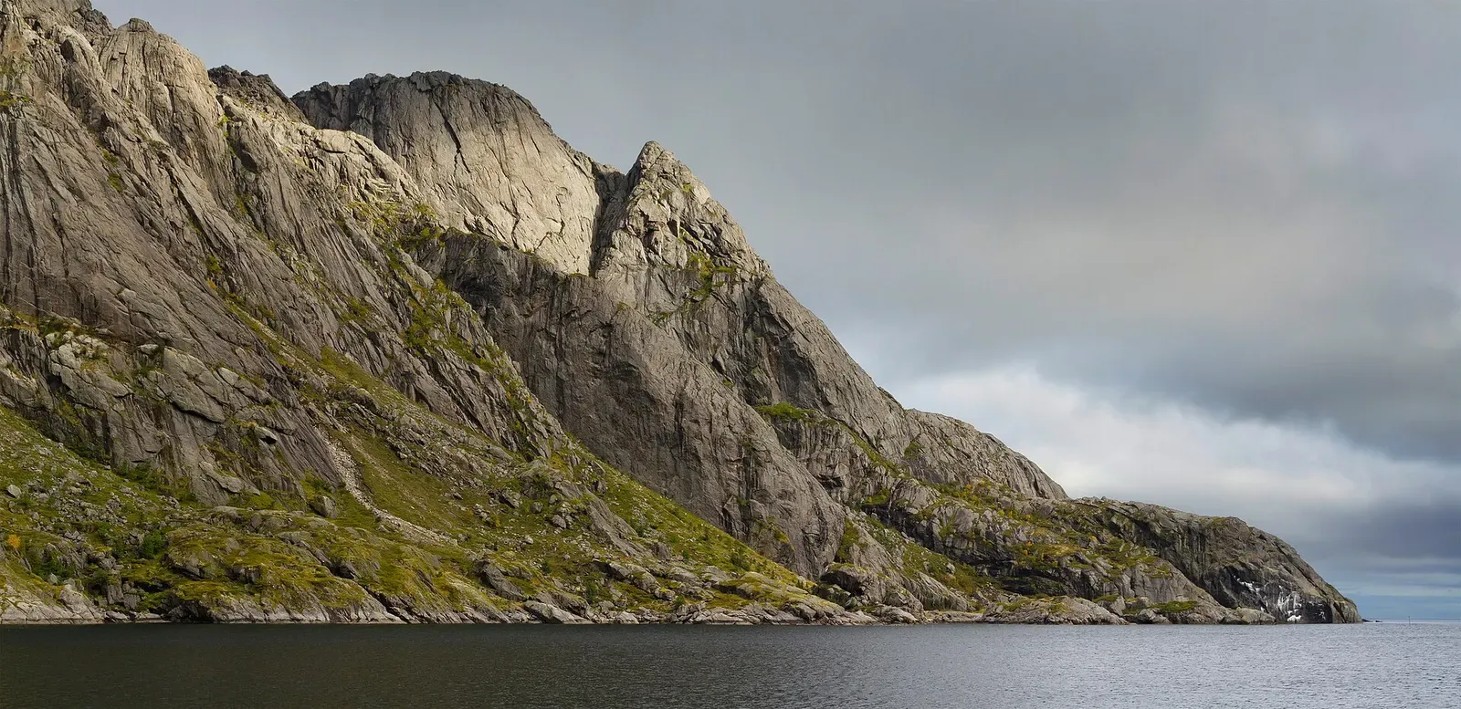 Steep bare granite ridge dropping straight into dark calm water under a grey sky — Shit Wall, Lofoten Islands