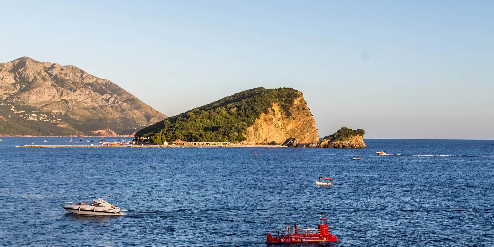 A green, cliff-edged island with a small rocky islet at its tip, seen across blue water with a white motorboat and a red boat in front — Galiola, Budva & Jaz
