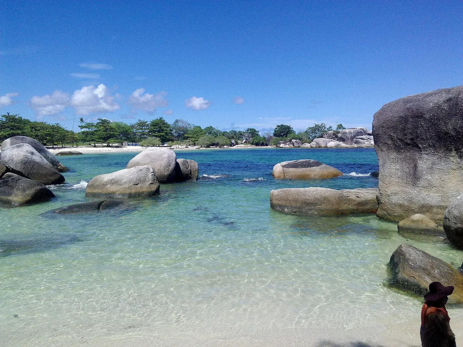 Clear turquoise shallows among huge rounded grey granite boulders, with a white sand beach and trees behind under a blue sky — Belitung