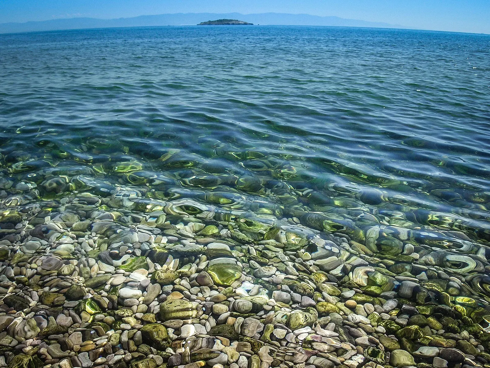 Clear turquoise water over a pebble shoreline with the small islet of Tavşan Adası visible on the horizon — Bodrum