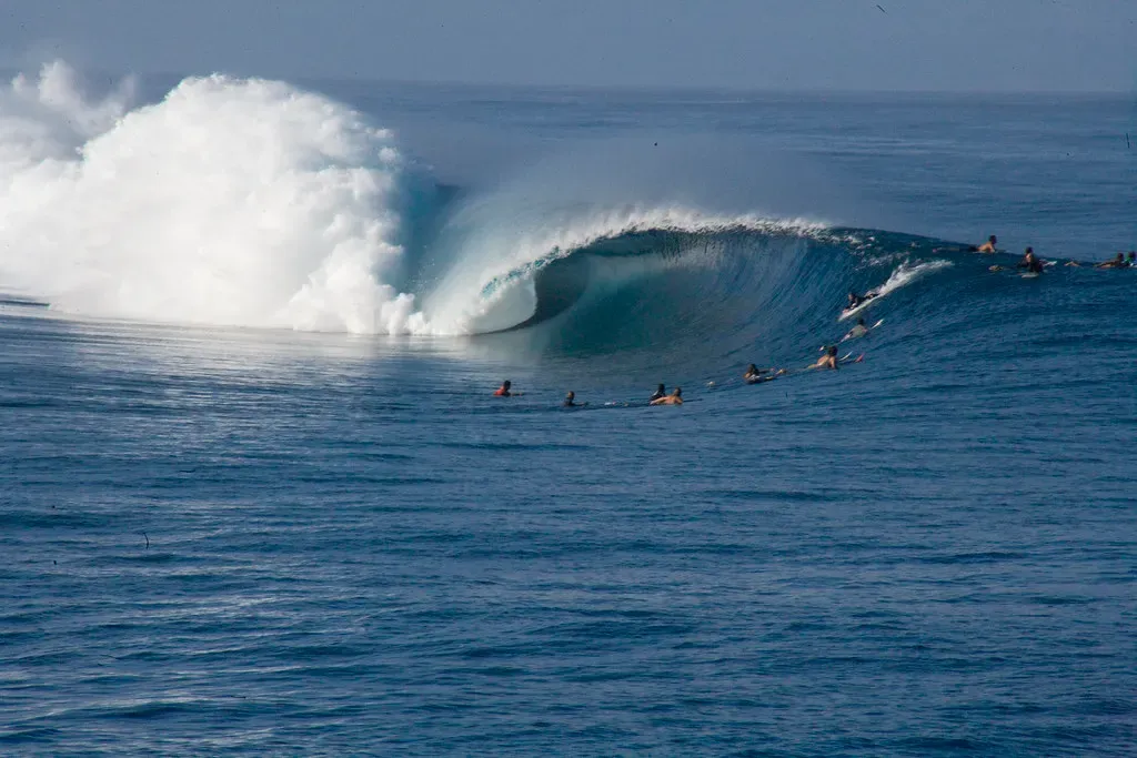 A deep blue wave curls into a hollow barrel and explodes into white spray while surfers on boards wait in the channel beside it — Tahiti & Teahupo'o.