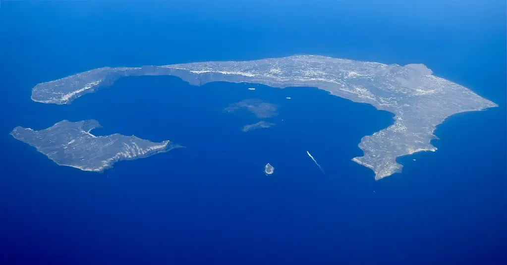 Aerial view of a ring of islands around a deep blue bay, with two small dark islets in the middle and a boat wake on the water — Santorini & the Caldera