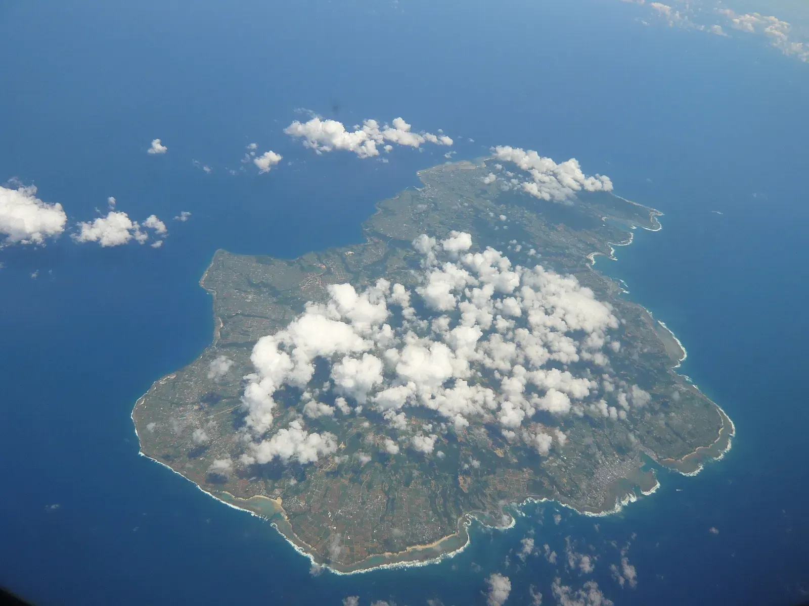 Aerial view of a green island ringed by a thin pale band of fringing reef in deep blue sea, with scattered white clouds over it — Tokunoshima