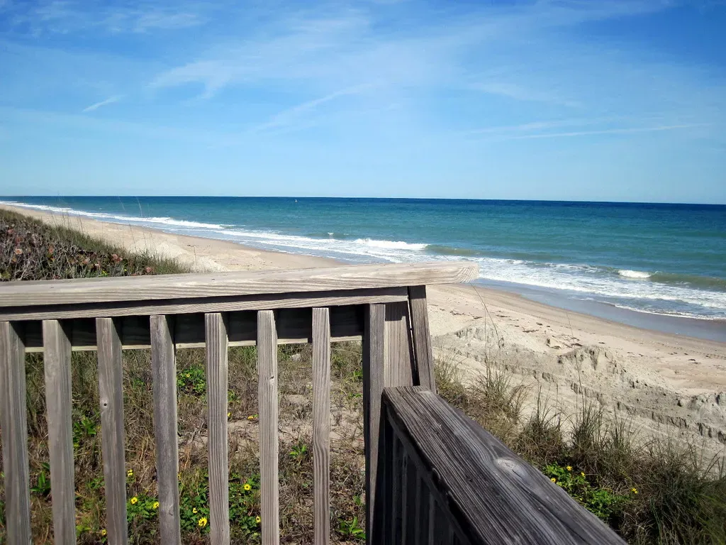 An empty sandy beach and blue Atlantic surf seen from a wooden dune walkover, with green dune plants below the railing — Treasure Coast.