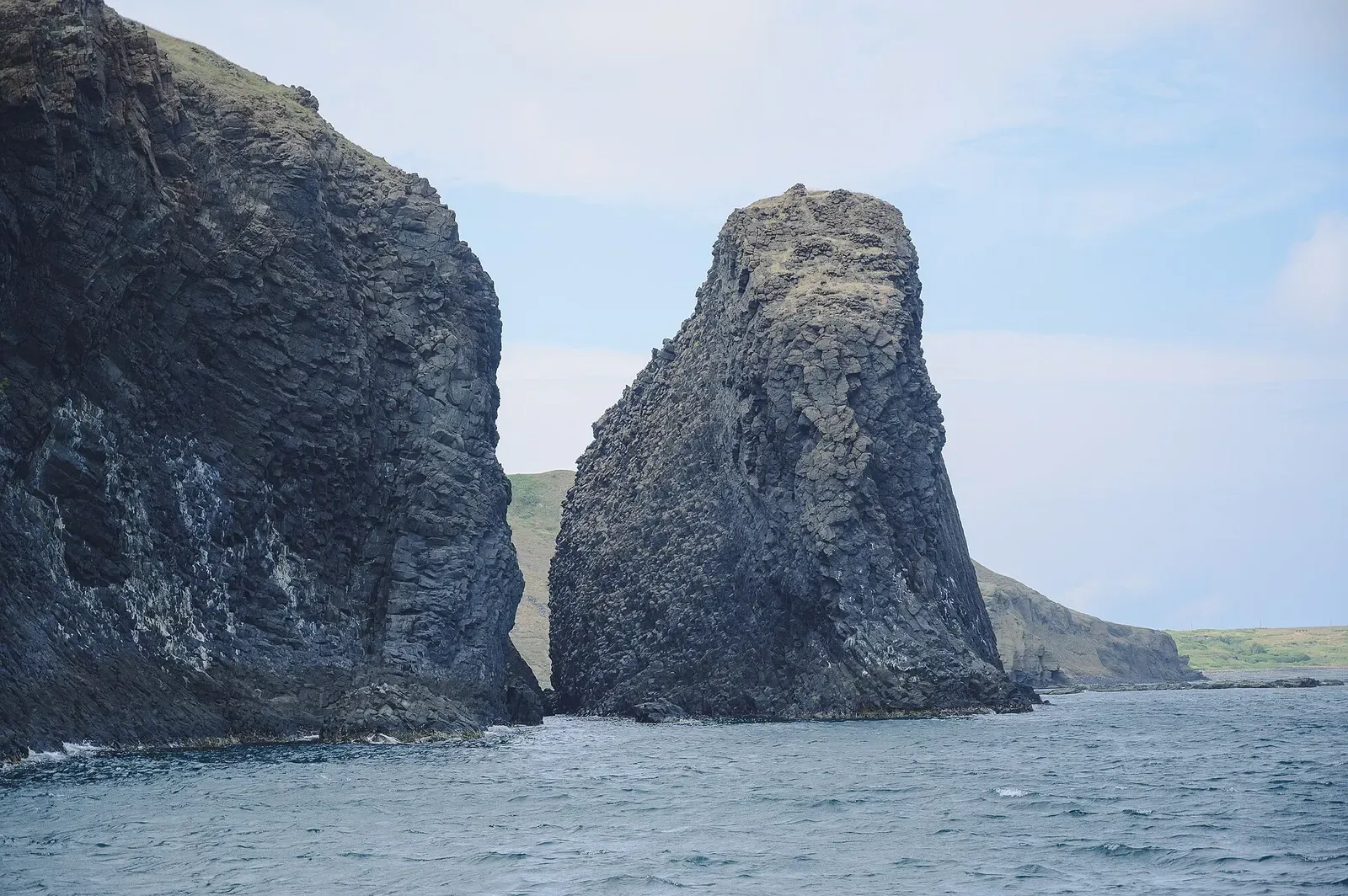 Two dark, jagged basalt sea stacks rising from grey-blue water, with a green headland behind under a pale sky — Penghu Islands