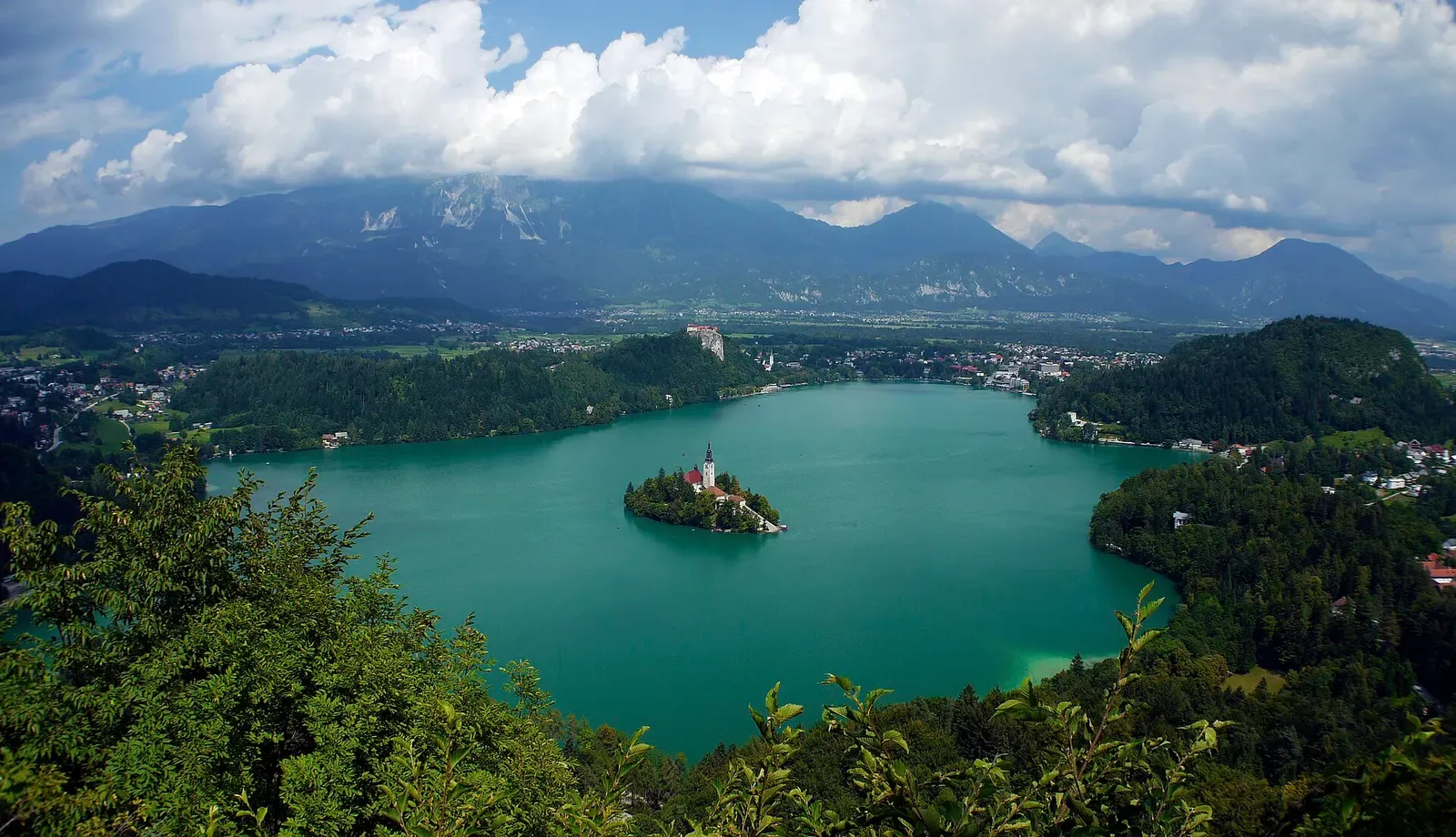 Lake Bled seen from a wooded hillside, green water around a small island with a church tower, mountains under cloud behind