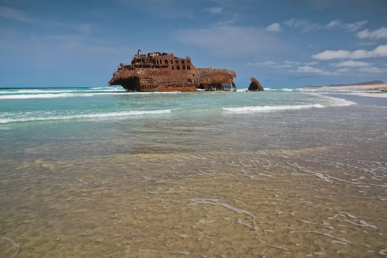 Rusted hull of a large wrecked ship sitting in shallow turquoise surf off a sandy beach under a blue sky — Boa Vista