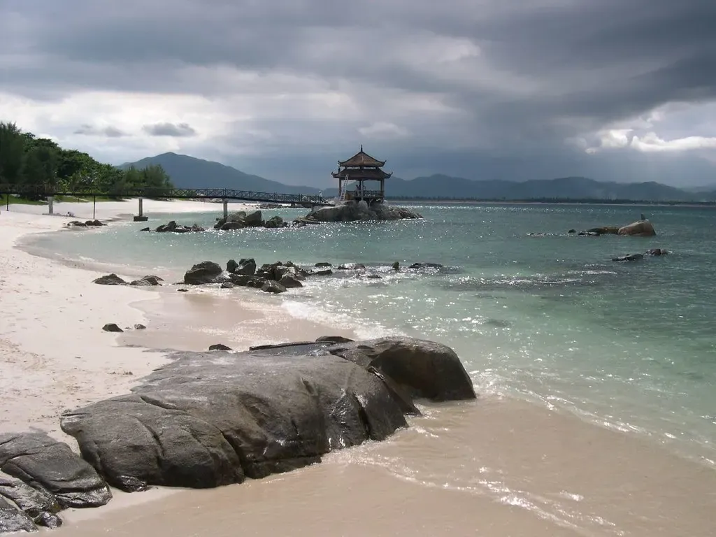 Dark boulders on a white sand beach, clear green shallows and a pavilion on a rock reached by a footbridge, under heavy storm clouds — Sanya & Wuzhizhou Island.