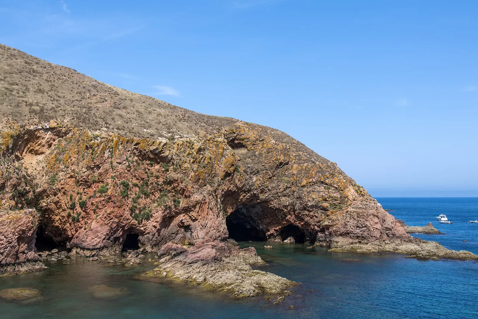 Sea caves cut into pink granite cliffs above clear blue water, with small boats offshore — Berlengas & Peniche
