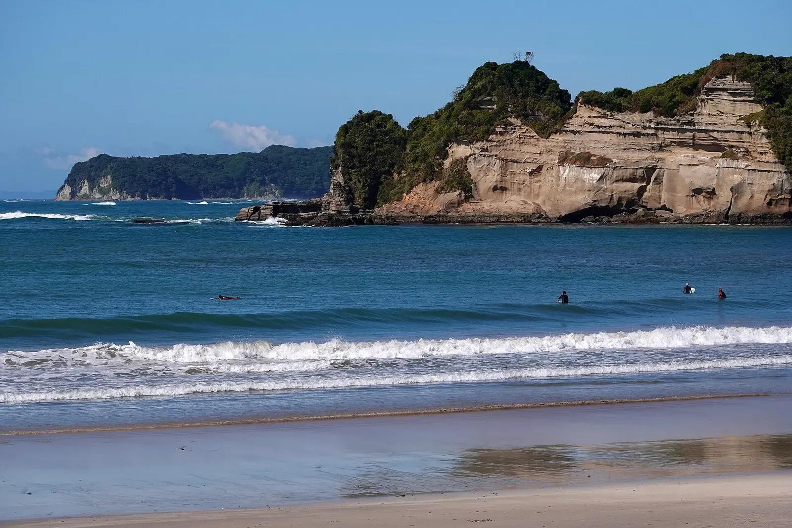 Waves rolling onto a sandy beach below pale layered sandstone cliffs topped with green trees, a few surfers in the water — Katsuura & Namegawa