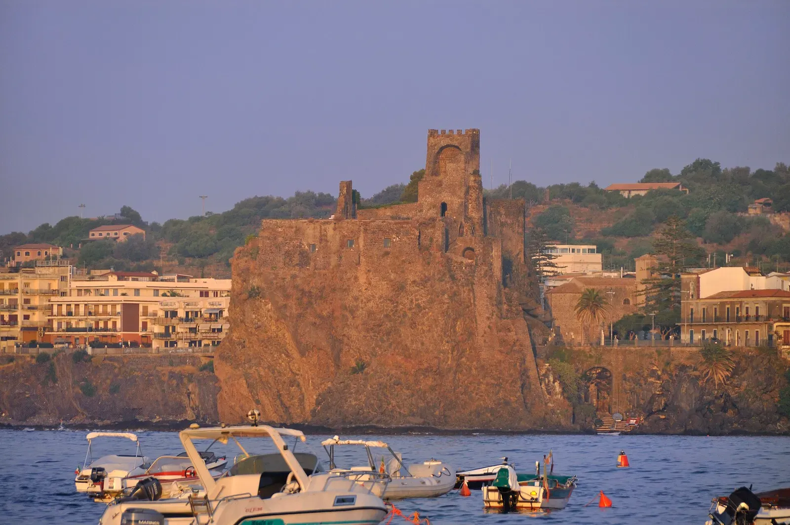 Aci Castello viewed from the water in golden evening light, boats moored below the rock