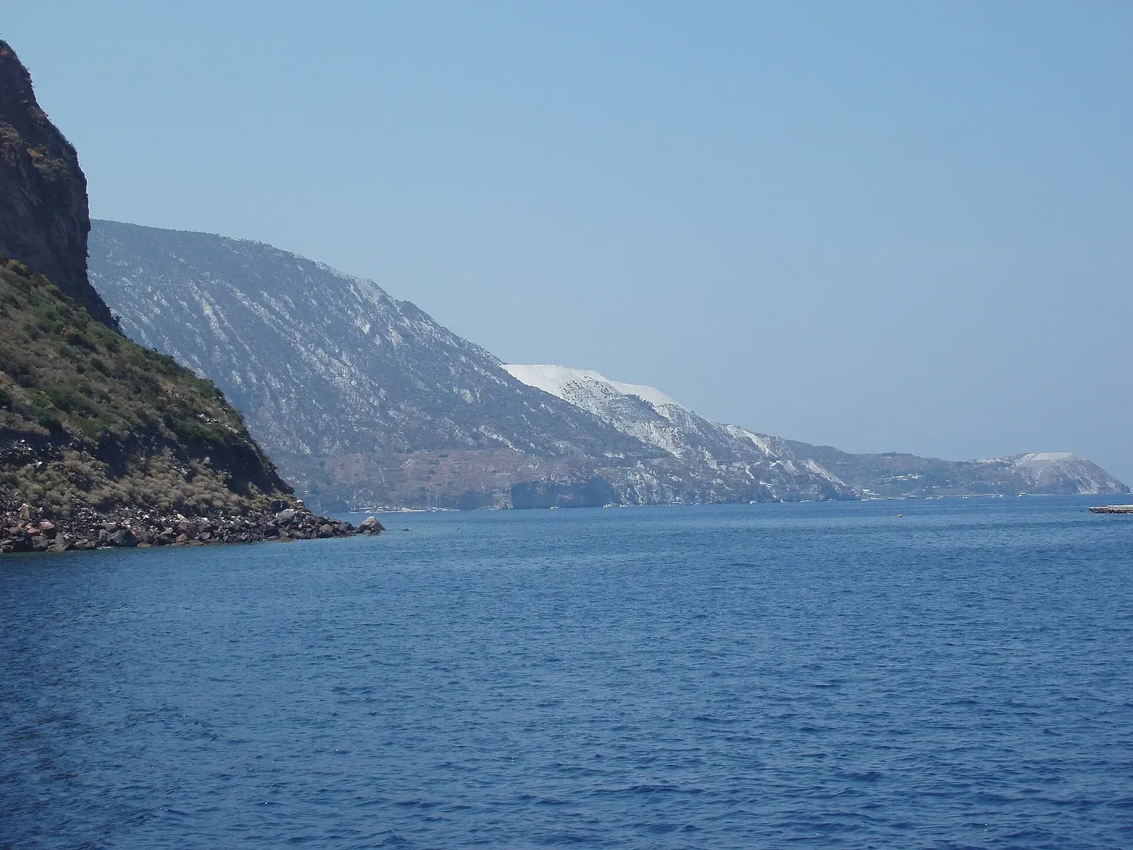 A steep island coast with white-streaked slopes running down to calm blue water, seen from a boat — Aeolian Islands