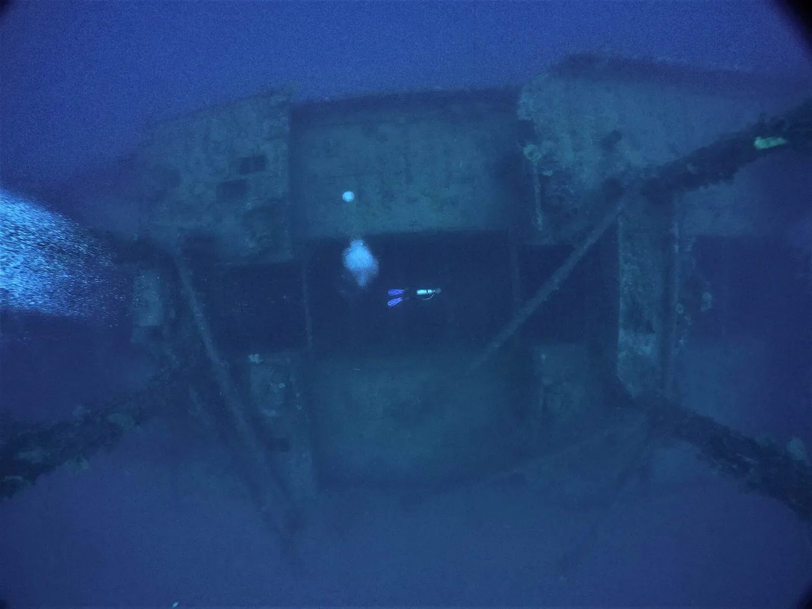 A diver with a torch hangs inside a dim blue wreck structure, framed by beams and openings — Aikoku Maru, Chuuk Lagoon.