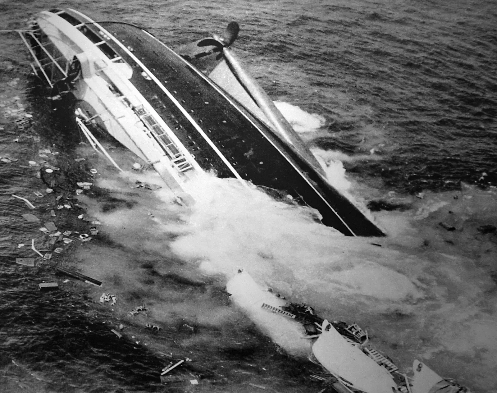 Aerial view of the Andrea Doria capsized and half-submerged, listing hard with debris scattered on the water.
