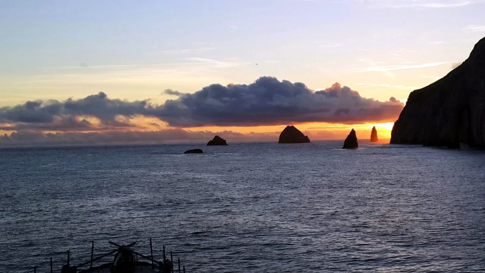 Row of jagged sea stacks silhouetted against an orange sunset beside a dark cliff, calm swell in the foreground — Malpelo