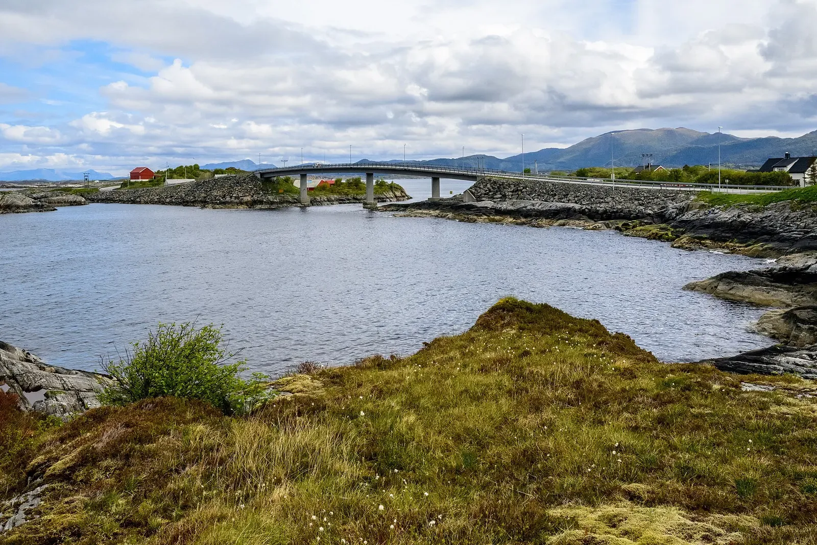 A sheltered bay ringed by rocks and grass, with a long low bridge, red houses and mountains on the far shore — Kristiansund & the Atlantic Ocean Road.