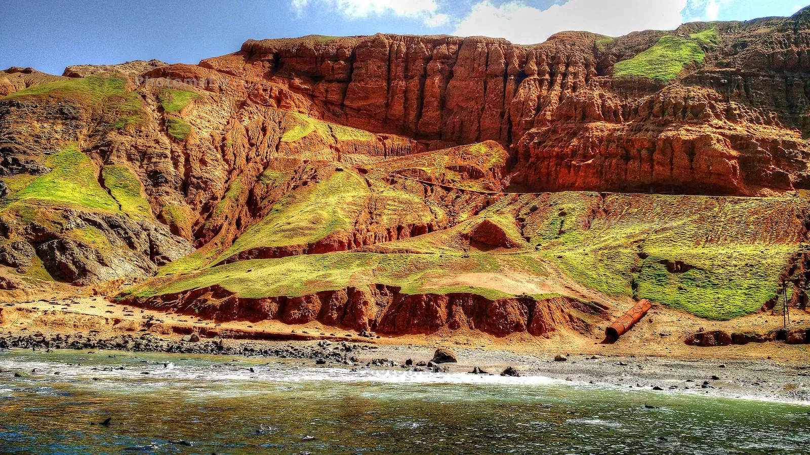 Eroded red and ochre volcanic cliffs streaked with green above a rocky shoreline at Bahía del Padre — Robinson Crusoe Island (Juan Fernández)