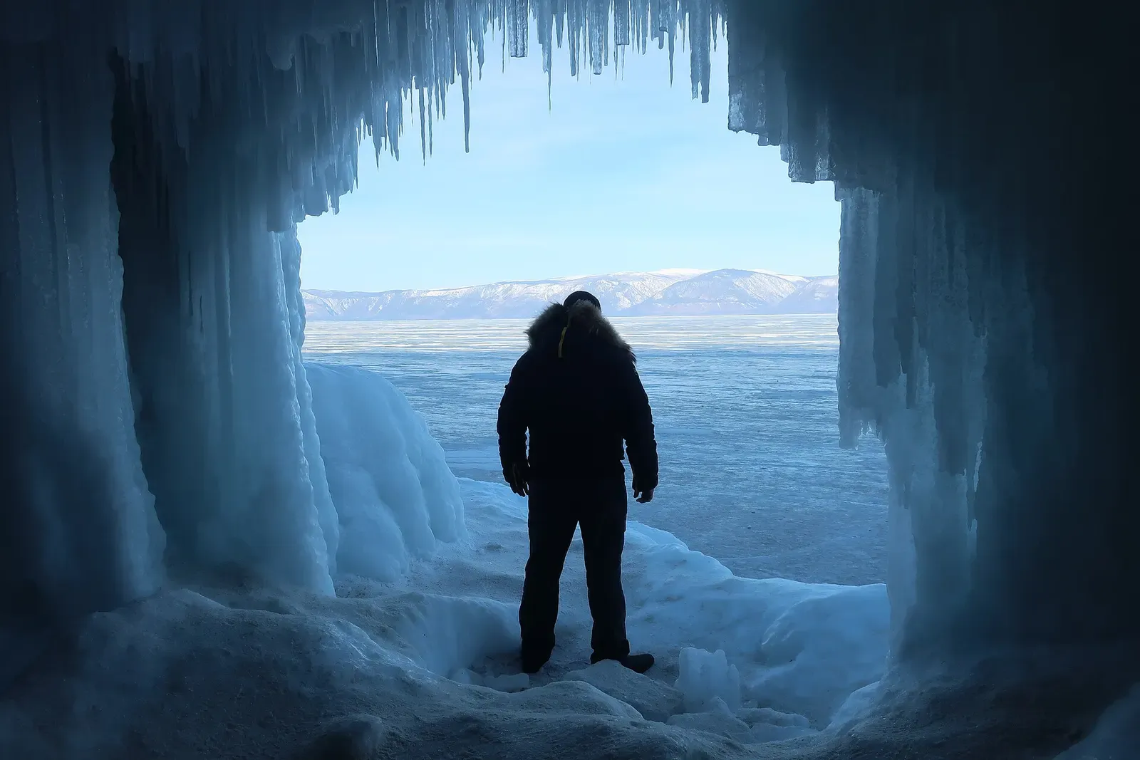 A person in a dark parka stands silhouetted inside an ice grotto hung with icicles, looking out over a frozen lake to snowy hills — Lake Baikal.