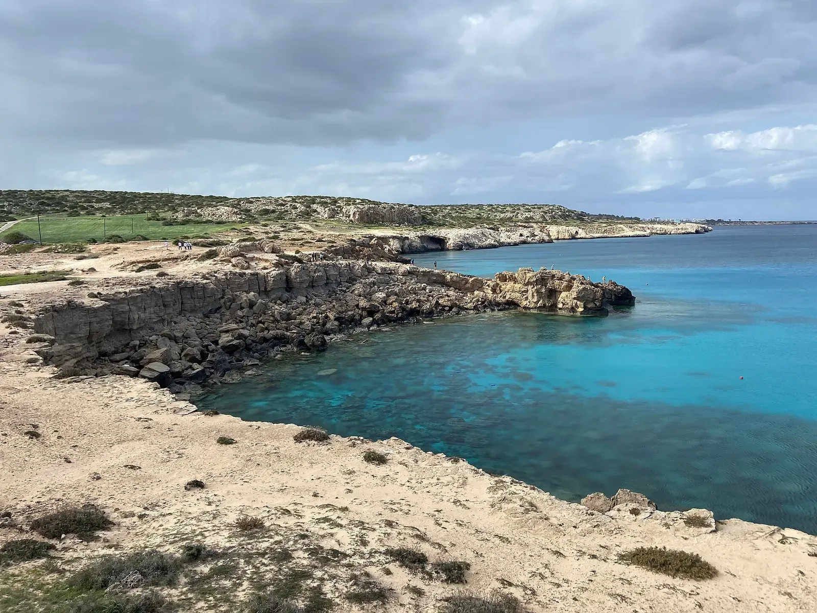 A low rocky shoreline enclosing a turquoise cove with dark patches on the shallow bottom, under a cloudy sky — Cape Greco & Ayia Napa