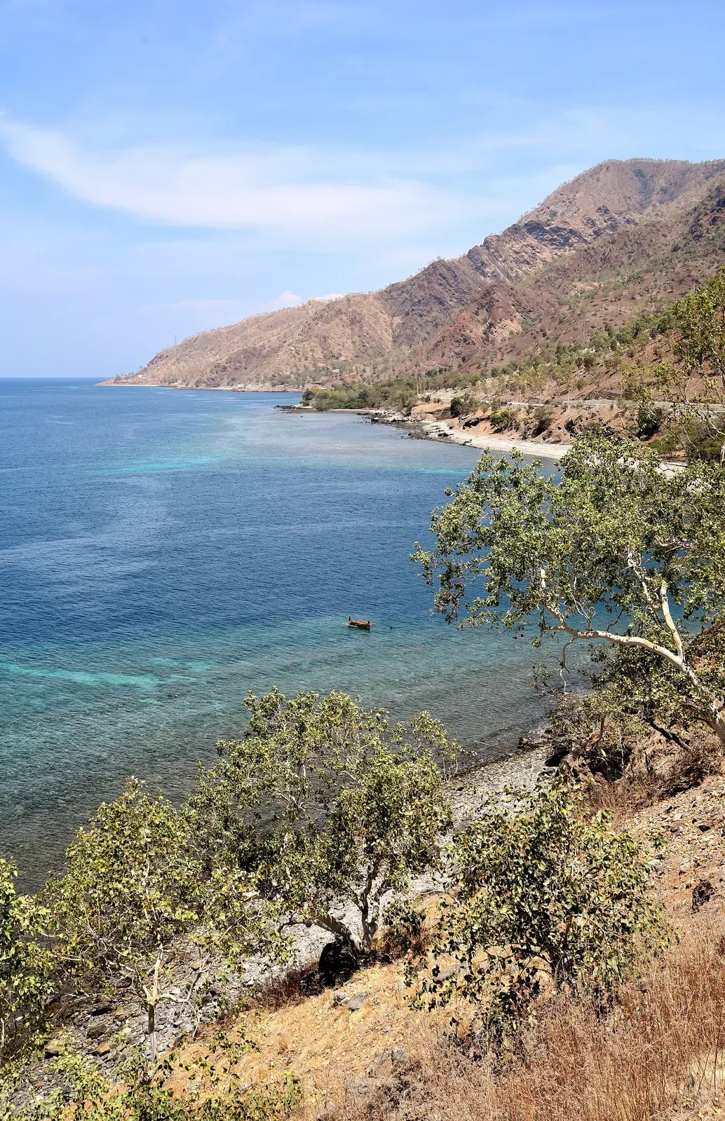 Turquoise fringing reef along a dry, steep coastline with a few small boats offshore — Bob's Rock, Dili