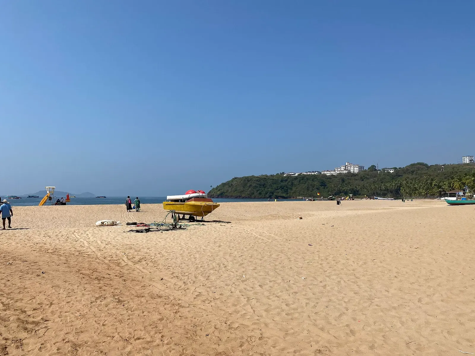 A yellow speedboat on an open sand beach with a wooded headland and calm sea behind, under a clear blue sky — Goa & the Grande Island
