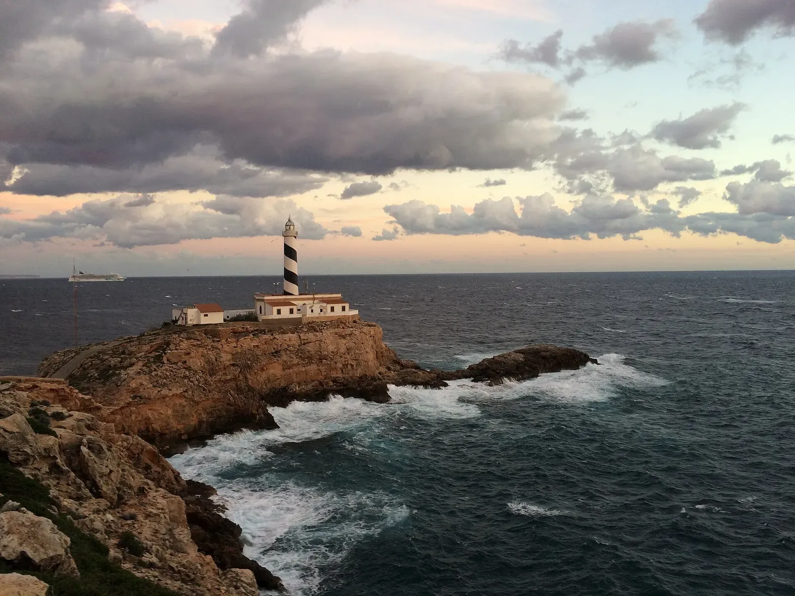 The Cap de Cala Figuera lighthouse on its rocky point at dusk with waves breaking below