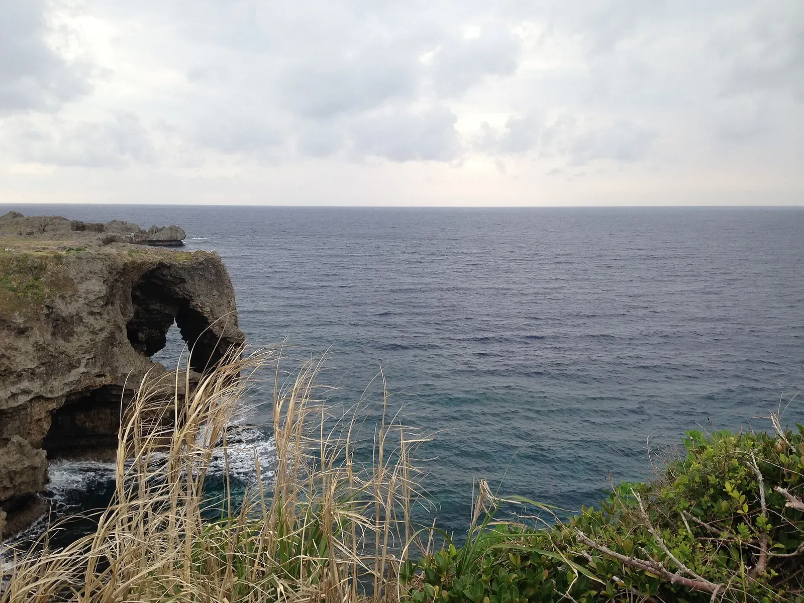Cape Manzamo's rock arch seen from the clifftop grass, overlooking open sea — Horseshoe Cliff, Okinawa.