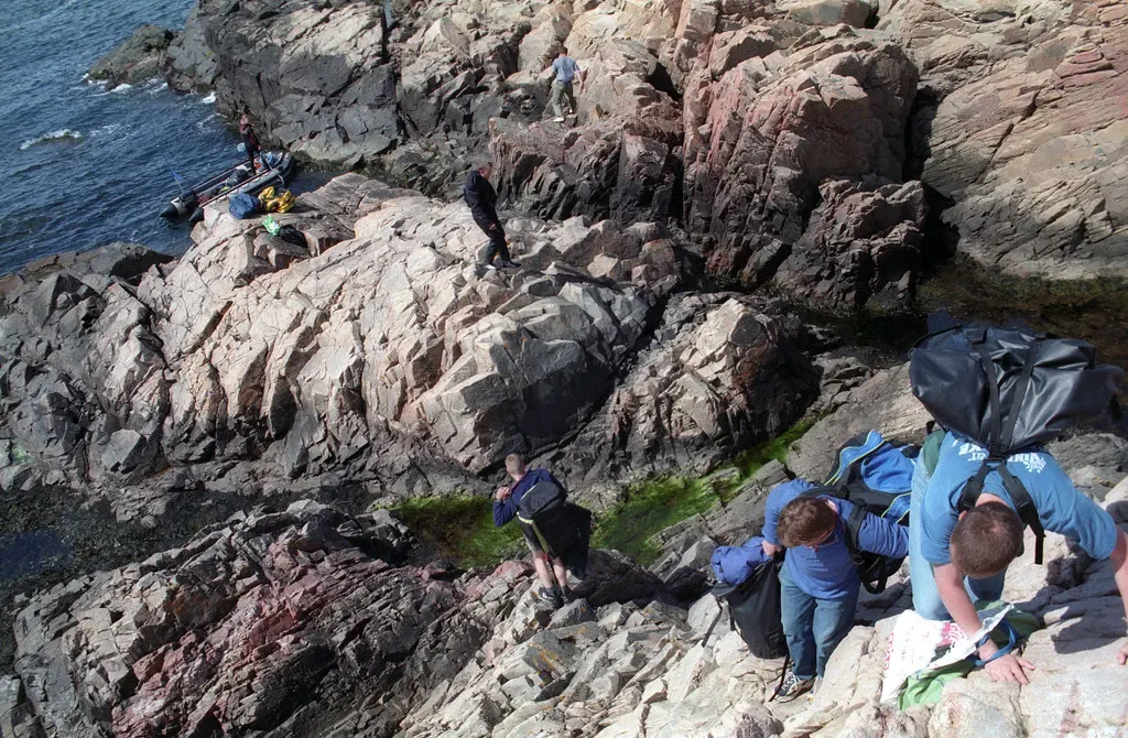 Divers carrying bags climb down steep pink and grey rocks toward the sea, where an inflatable boat waits by the shore — Kullen, Kullen, Sweden.