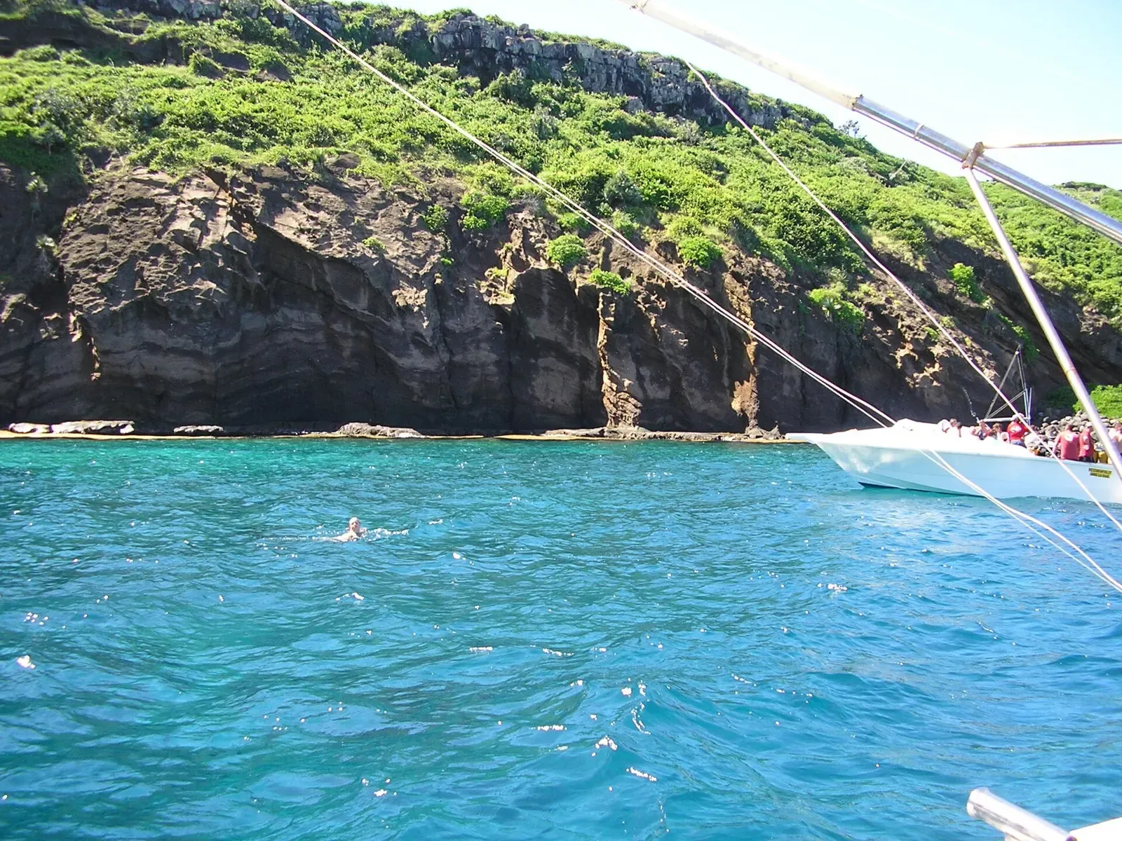 A swimmer in clear turquoise water beside a sailing boat under the cliffs of Ile Coin de Mire — Djabeda, Mauritius.