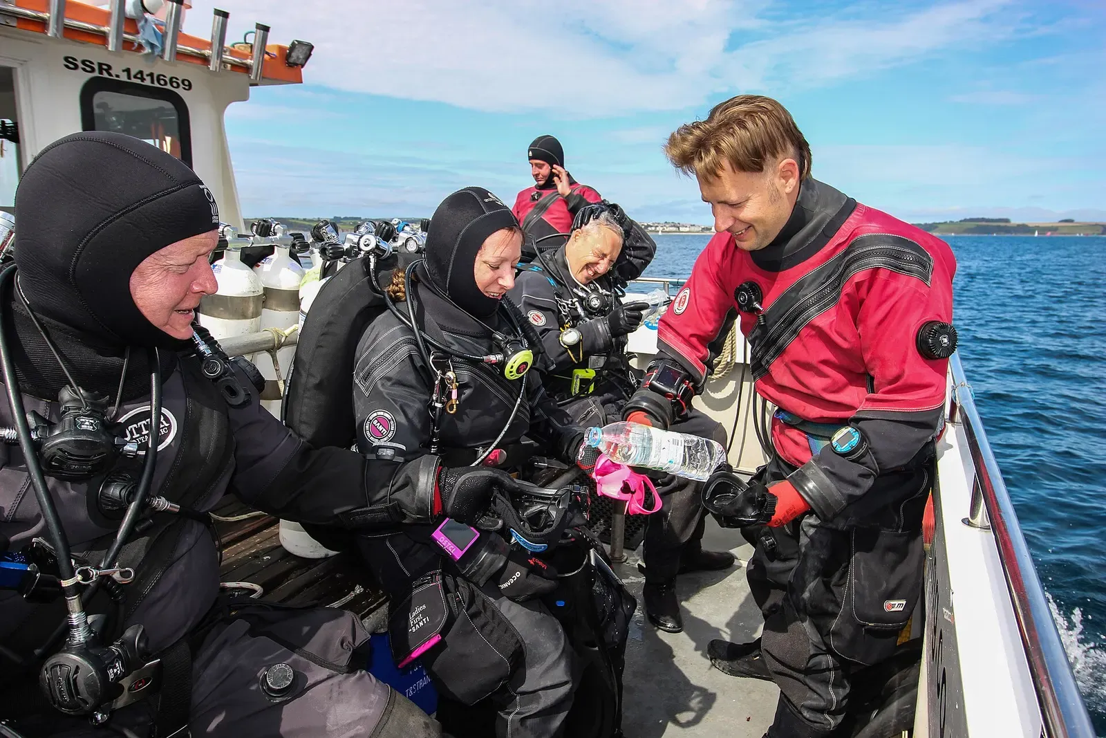 Four divers in drysuits and hoods on a boat deck lined with cylinders, one in a red drysuit holding a bottle — Cornwall