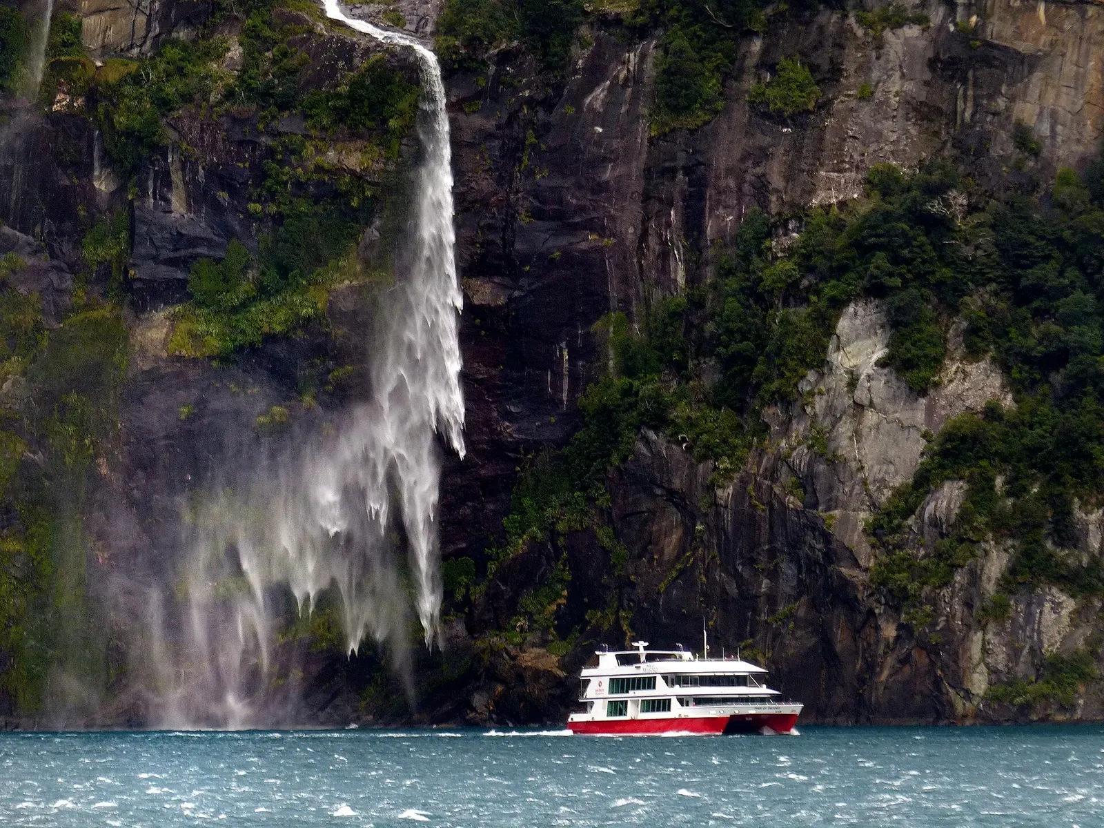 A waterfall dropping down the cliffs of Milford Sound past a small tour boat — Fiordland