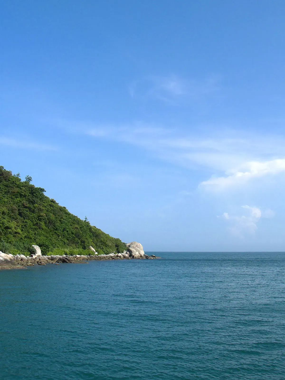 Jungle-covered headland ending in pale granite boulders at the waterline under a blue sky — Cham Islands & Hoi An