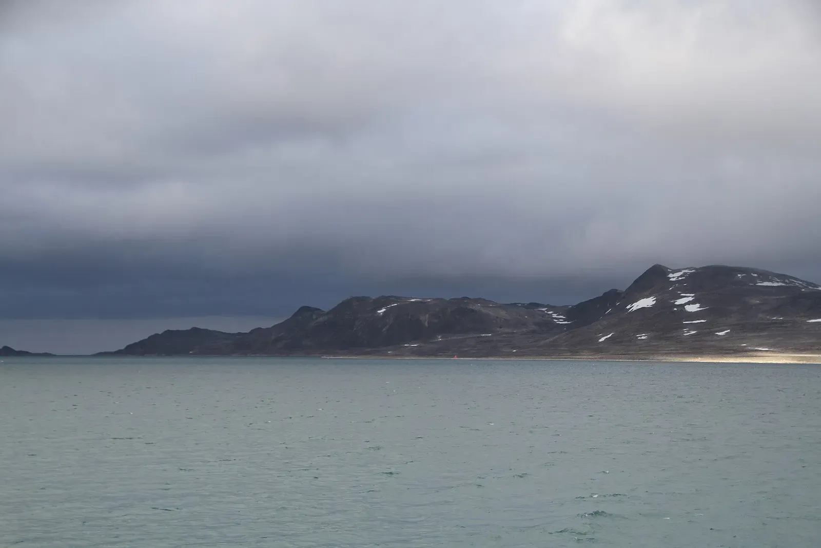 Dark mountain ridge above the grey water of Smeerenburgfjorden, seen from Danskøya to the southeast — Svalbard.
