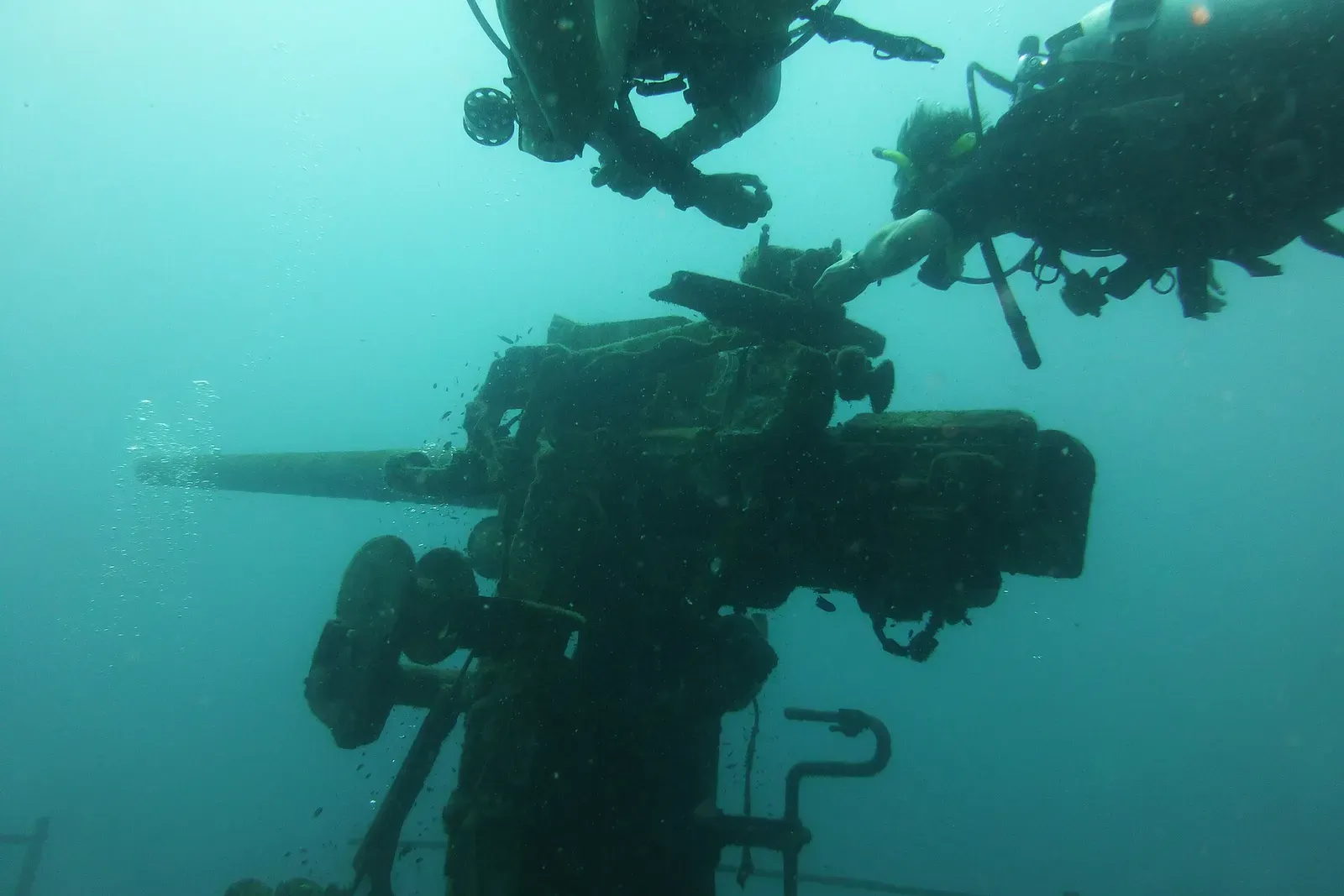 Diver silhouetted above the wreck's gun mount in green water — HTMS Sattakut, Koh Tao