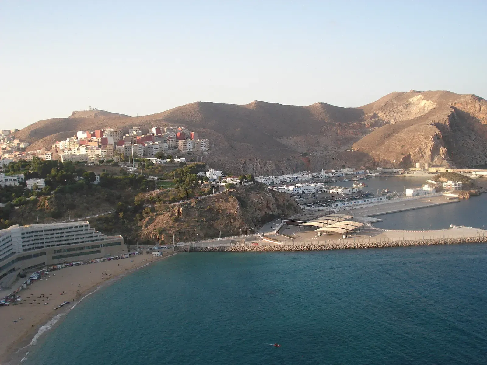 Curving beach and the harbour breakwaters of Al Hoceima under brown mountains, seen from a hillside