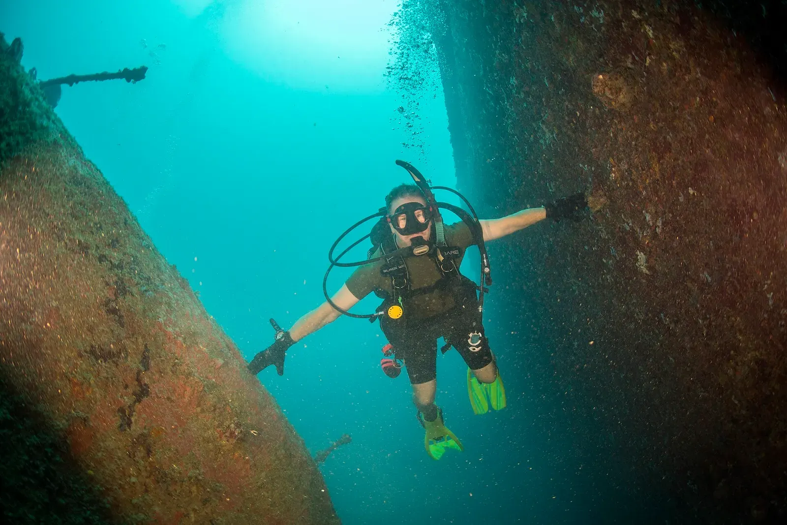 A diver ascends between two rust-covered hulls with a gun barrel visible in the background — SMS Cormoran & Tokai Maru, Guam