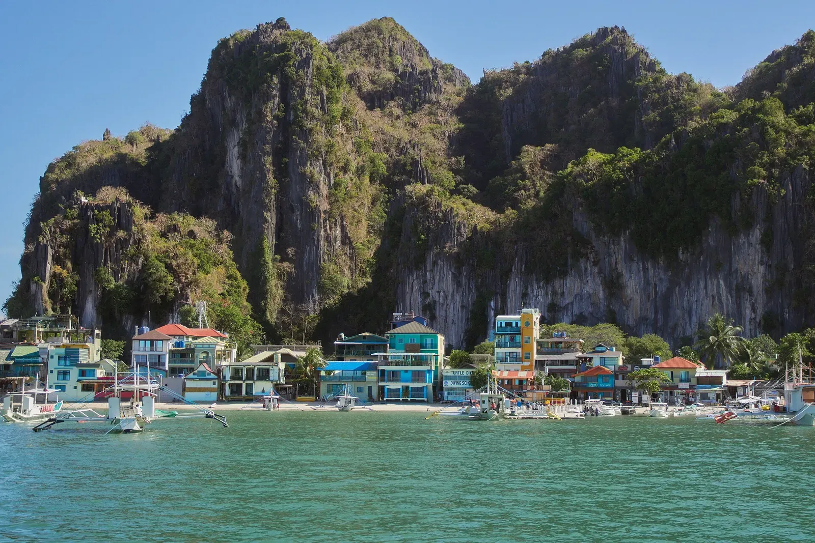 El Nido town's waterfront buildings and moored boats beneath tall limestone karst cliffs