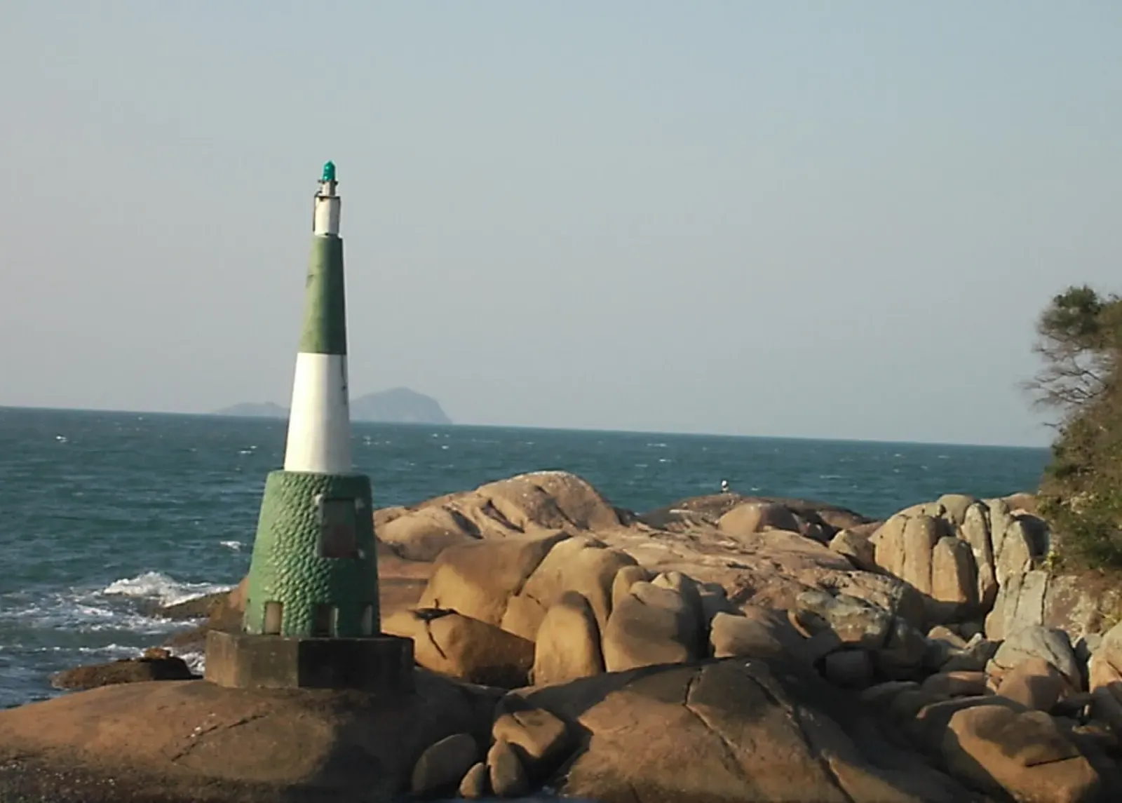 Small green and white light tower on granite boulders, with open sea and an island on the horizon — Florianópolis & Ilha do Xavier