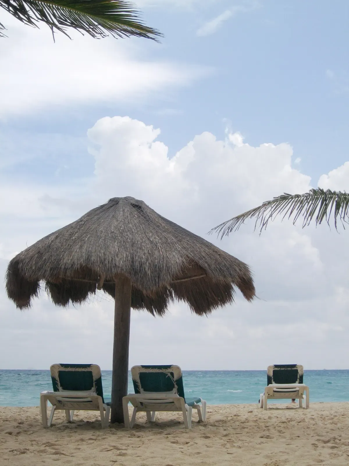 A thatched palapa umbrella shades empty beach loungers on the white sand of Playa del Carmen, with turquoise Caribbean water beyond