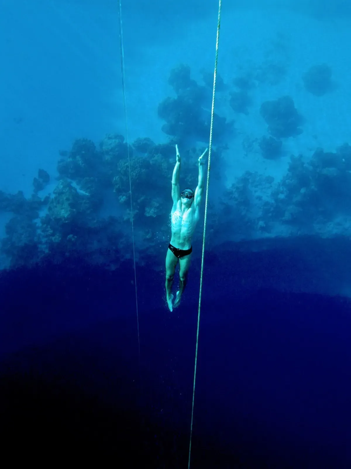 A freediver descends headfirst along a guide rope into the deep blue interior of Dean's Blue Hole, rock formations visible on the walls below