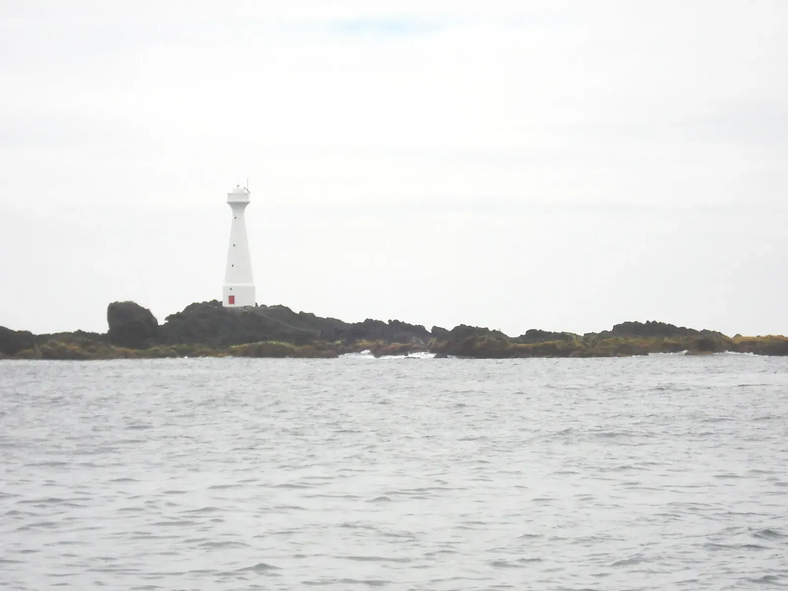 A white lighthouse stands on a low, dark rocky islet across a grey sea under an overcast sky — Ilhéus das Formigas, Azores